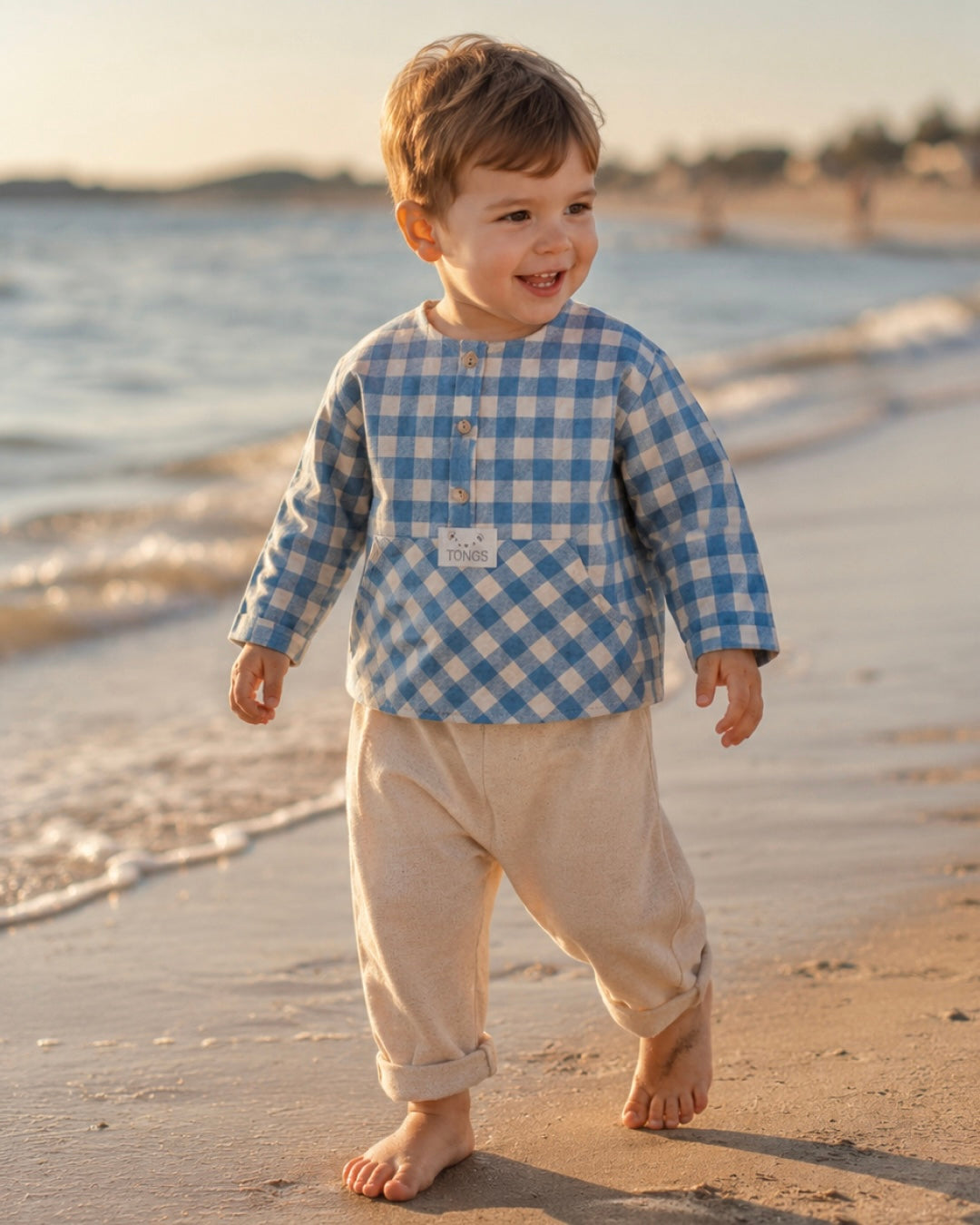 Baby boy in blue plaid cotton outfit set with pants walking on the beach